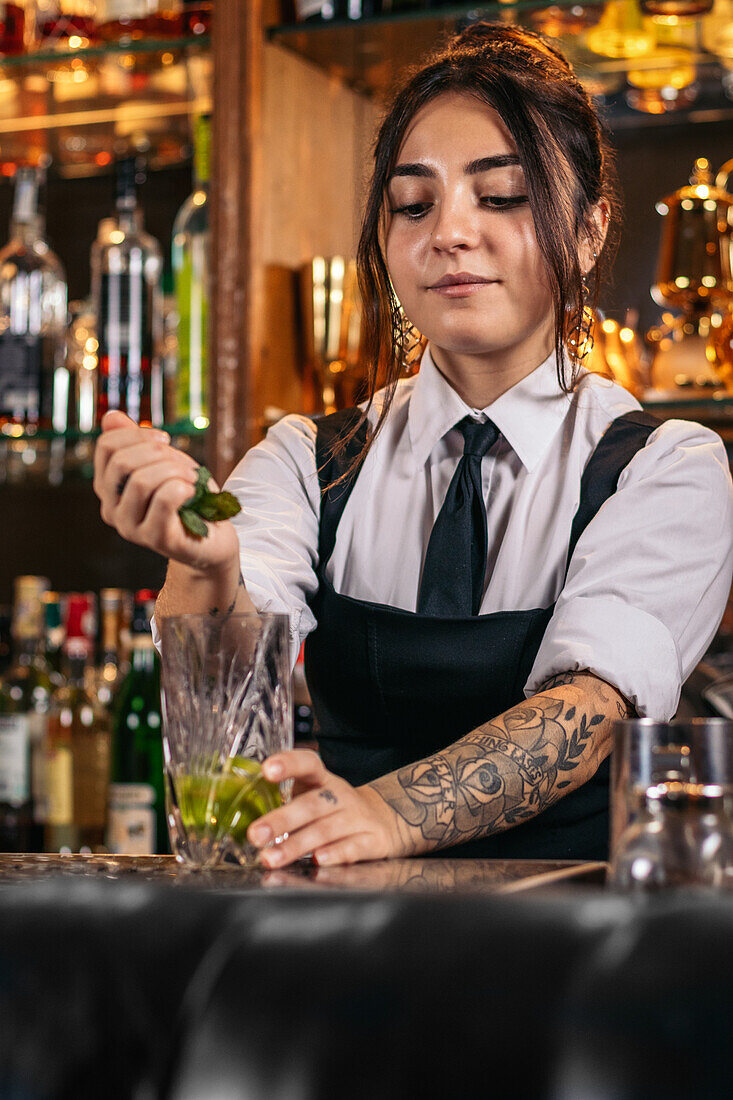 Bartender adding fresh mint leaves in glass while preparing Mojito cocktail at counter in bar