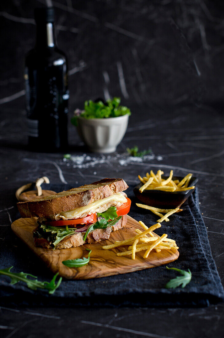 Leckeres Sandwich mit Tomate, Zwiebel, Käse und Fleisch auf einem Holzbrett mit Pommes frites und Messer auf einem blauen Tisch