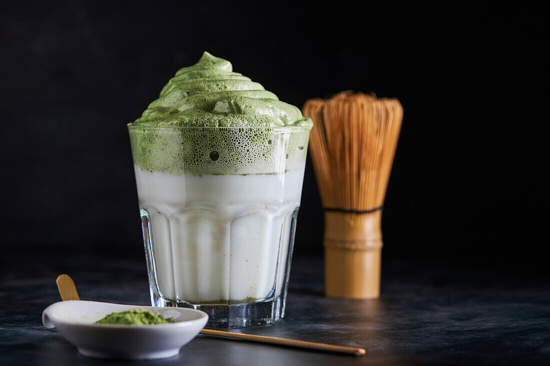 Glass of fresh dalgona coffee with matcha and Japanese tea ceremony accessories consisting of bamboo whisk called chasen and traditional scoop named chashaku placed on gray surface