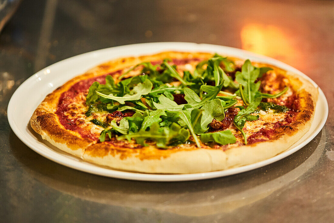 Leckere Pizza mit Parmesan und Pesto mit getrockneten Tomaten und frischem Rucola auf einem Teller auf einem Restauranttisch
