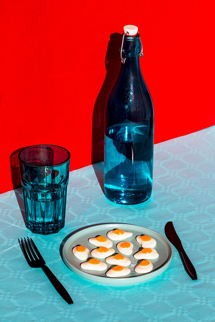 From above of composition of bottle of water with glass opposite plate with halved boiled eggs and fork and knife placed on blue surface against red background