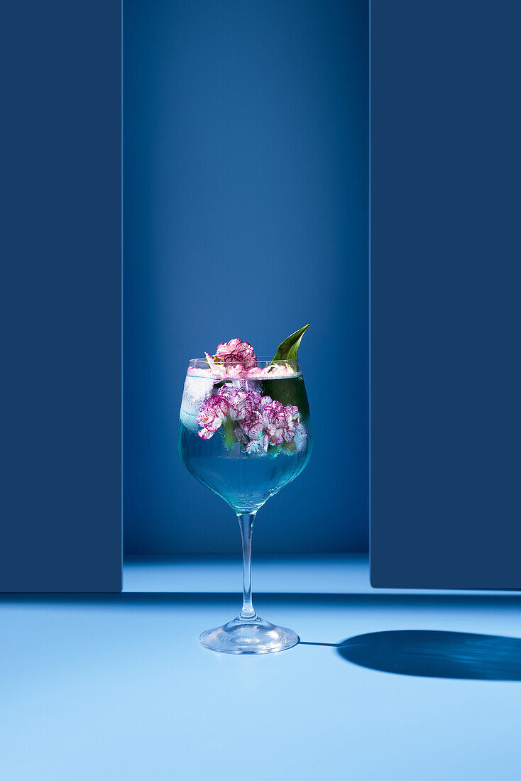 Transparentes Glas mit frischem Cocktail, Minzblättern und Blumen vor blauem Hintergrund auf dem Tisch