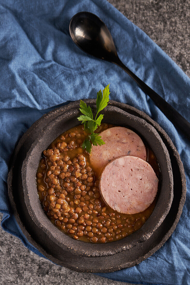 Rustic bowl with tasty lentil soup with parsley and slices of sausage placed on marble table and blue napkin during lunch