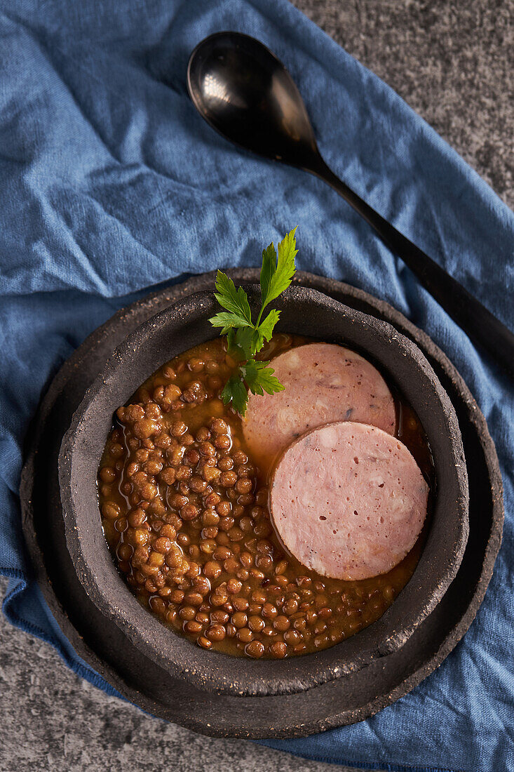 Rustikale Schale mit schmackhafter Linsensuppe mit Petersilie und Wurstscheiben auf Marmortisch und blauer Serviette beim Mittagessen