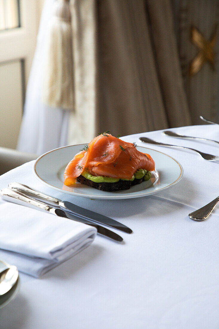 Blick von oben auf einen Teller mit leckerem Avocado-Toast mit pochiertem Ei und geräuchertem Lachs, serviert auf einem Tisch mit Besteck und Kaffeetasse während des Frühstücks im Hotelrestaurant