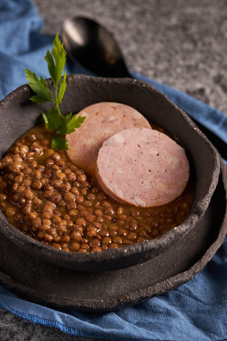 Rustikale Schale mit schmackhafter Linsensuppe mit Petersilie und Wurstscheiben auf Marmortisch und blauer Serviette beim Mittagessen