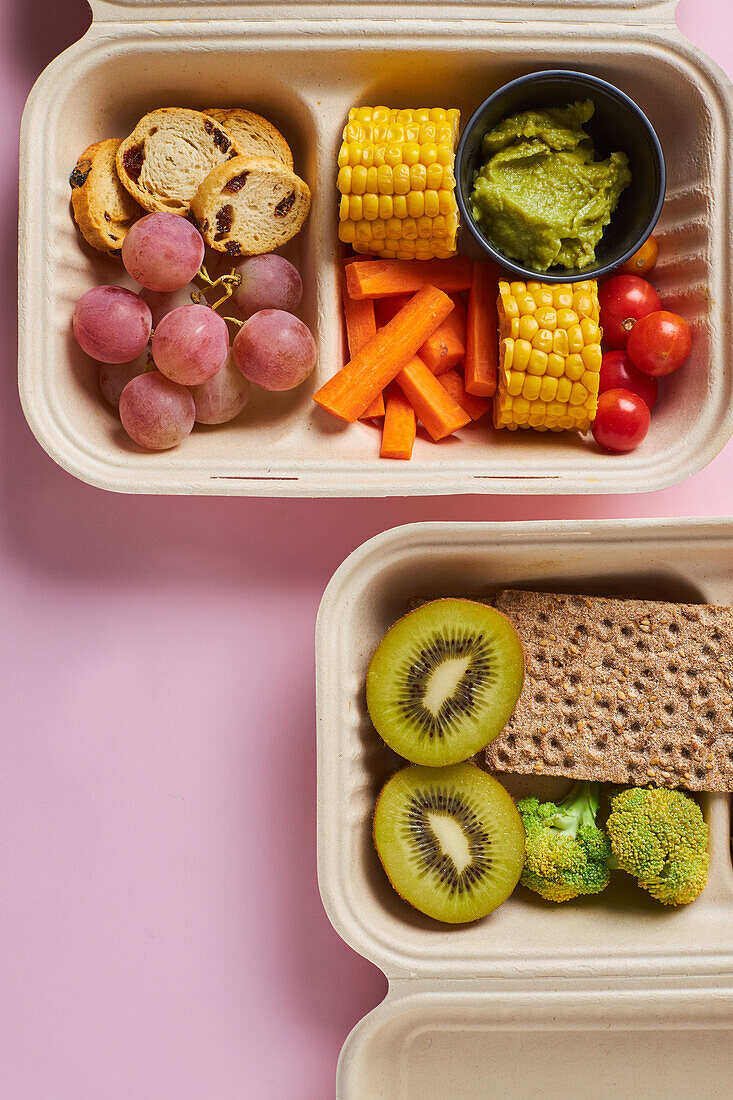 Von oben von Lunch-Boxen mit gesunden Lebensmitteln einschließlich Cracker Karotten-Sticks Trauben Kirschtomaten mit Kiwi Brokkoli Walnuss und Mandarine auf rosa Hintergrund