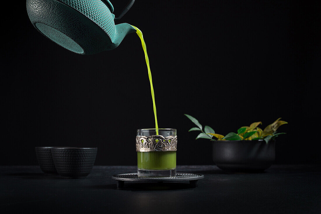 Gesunder japanischer Matcha-Tee, der während einer Teezeremonie aus einer grünen Teekanne in eine Glastasse mit Metallornamenten gegossen wird, vor schwarzem Hintergrund