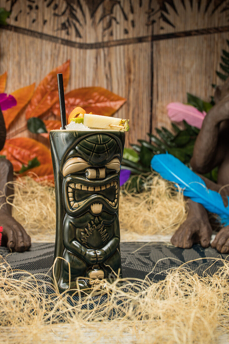 Large sculptural tiki cup filled with booze decorated with straw and fruits placed on green rug against dry grass