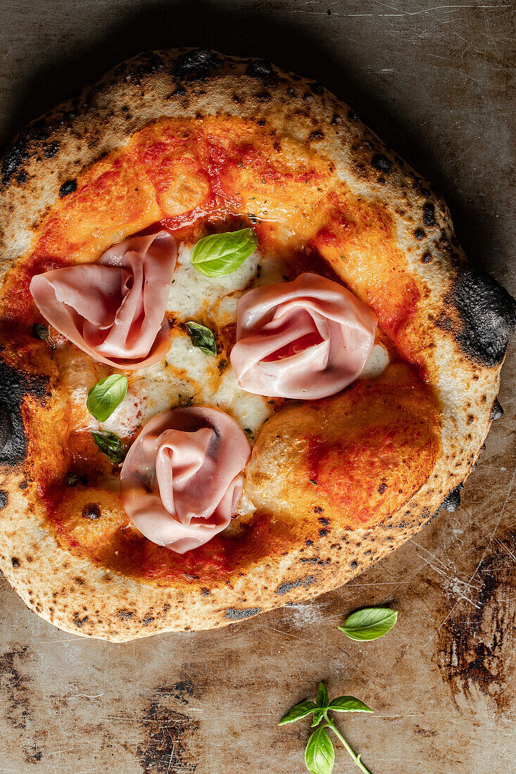 Top view of delicious pizza with cheese and ham and basil placed on burnt surface