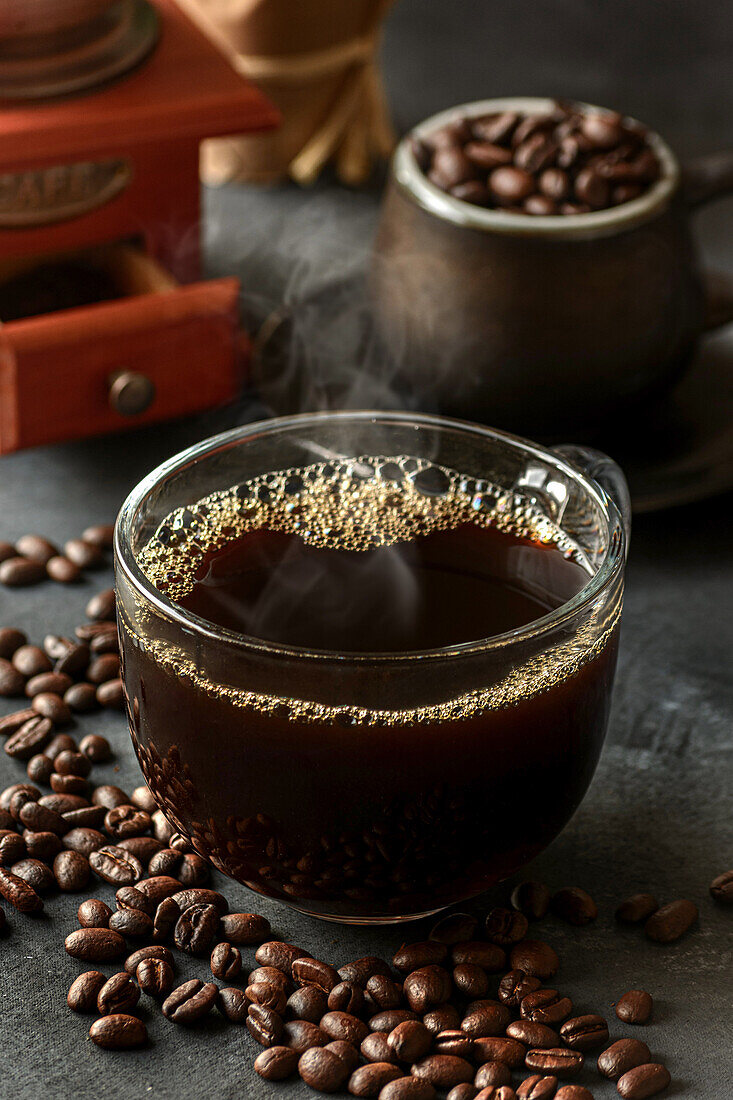 Glass of freshly brewed aromatic coffee on saucer placed on table with scattered beans against manual grinder