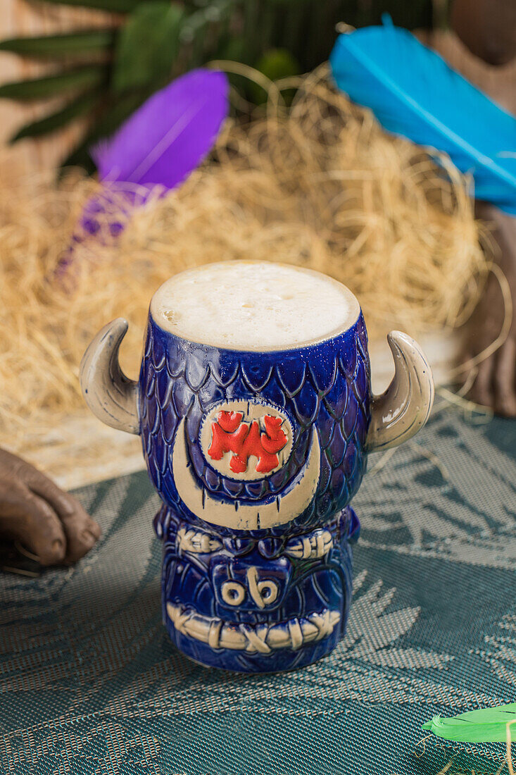 Stierförmiger Tiki-Becher mit alkoholischem Getränk mit Schaum vor trockenem Gras und Federn auf unscharfem Hintergrund von oben