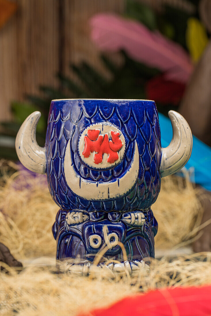 Stierförmiger Tiki-Becher mit alkoholischem Getränk und Schaum vor trockenem Gras und Federn im unscharfen Hintergrund