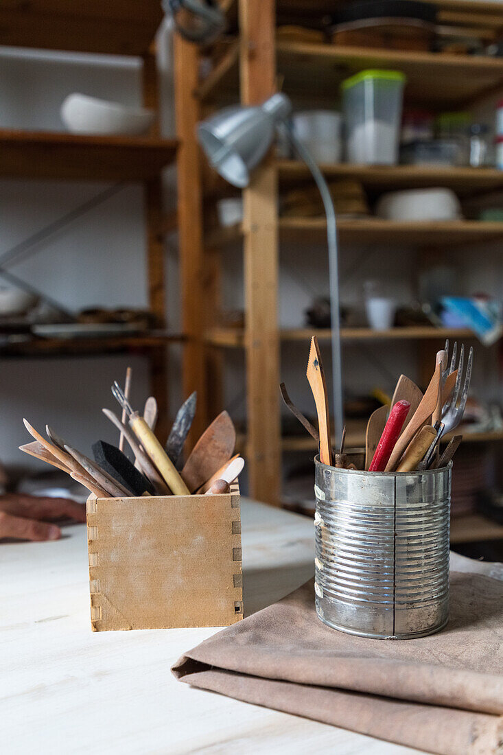 Various pottery tools in metal can and stationery holder placed on table in aged workshop with wooden shelves