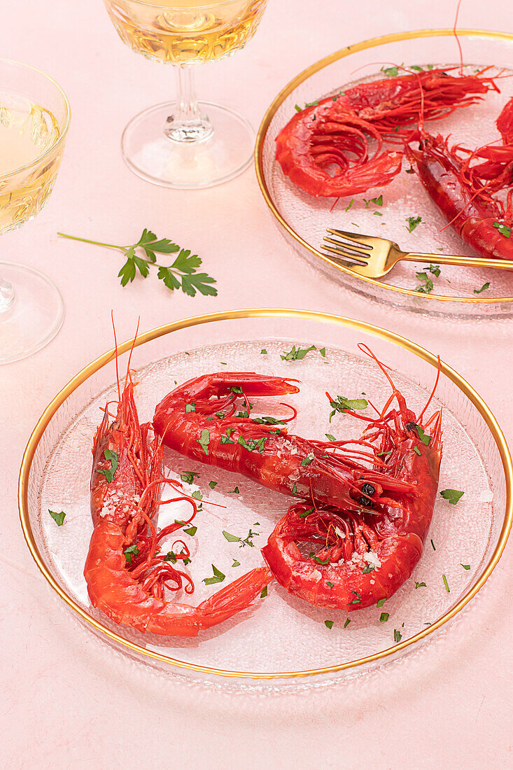Blick von oben auf einen Glasteller mit leckeren gebratenen Tiger Shrimps neben einem Glas Wein auf einem Tisch mit rosa Hintergrund