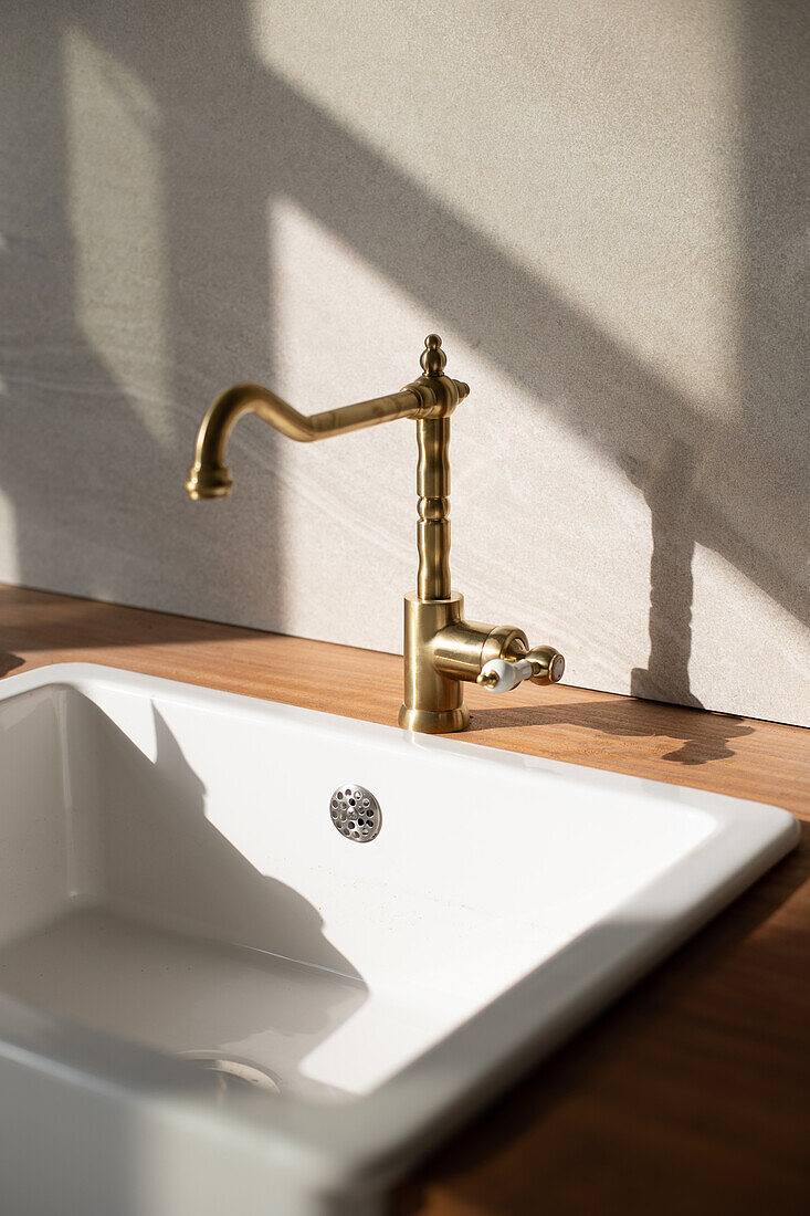 Holzschrank mit weißer Keramikspüle und goldenem Wasserhahn an der Wand in einer Küche mit hellem Sonnenlicht zu Hause