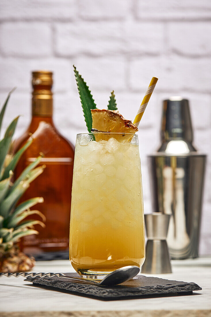 Yellow cocktail in glass garnished with pineapple piece and green leaves with paper straw placed on slate coaster with bar spoon