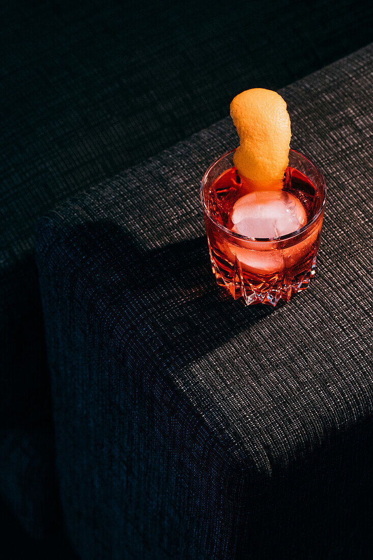 Glas erfrischender Negroni-Cocktail mit bitterem Geschmack und Eis, garniert mit Orangenschalen, serviert auf dem Arm der Couch in einem dunklen Raum
