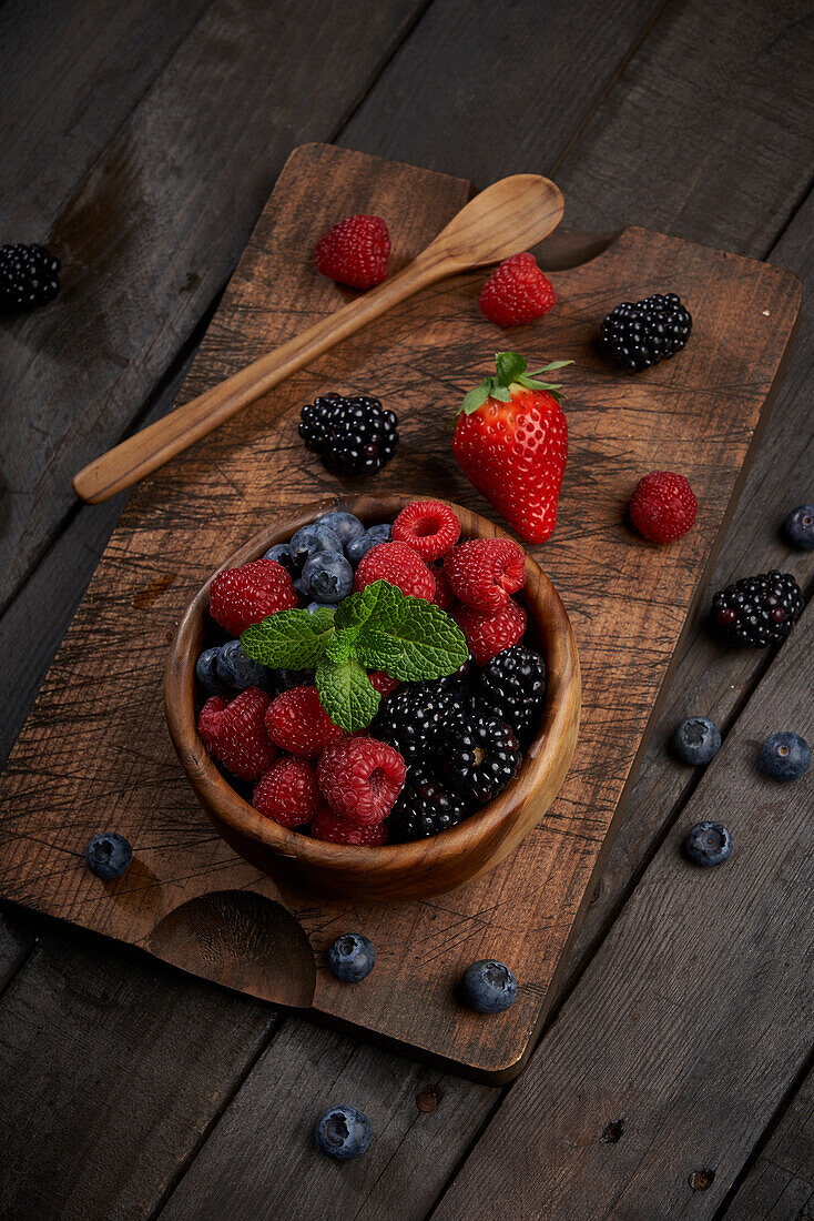 From above bowl with various ripe berries and mint leaves on wooden cutting board in light kitchen