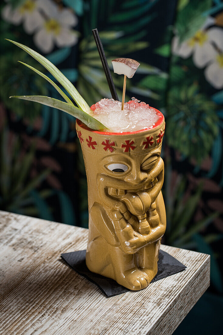 Polynesischer Tiki-Becher mit kaltem alkoholischem Getränk, dekoriert mit Strohhalm und grünen Ananasblättern, auf einem Holztisch