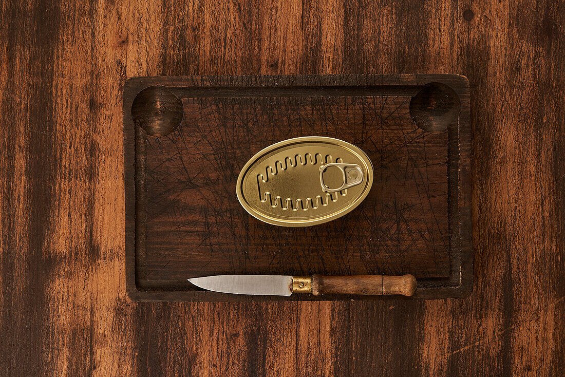 From above scratched chopping board with sharp knife placed near sealed can with preserved food on rustic lumber table