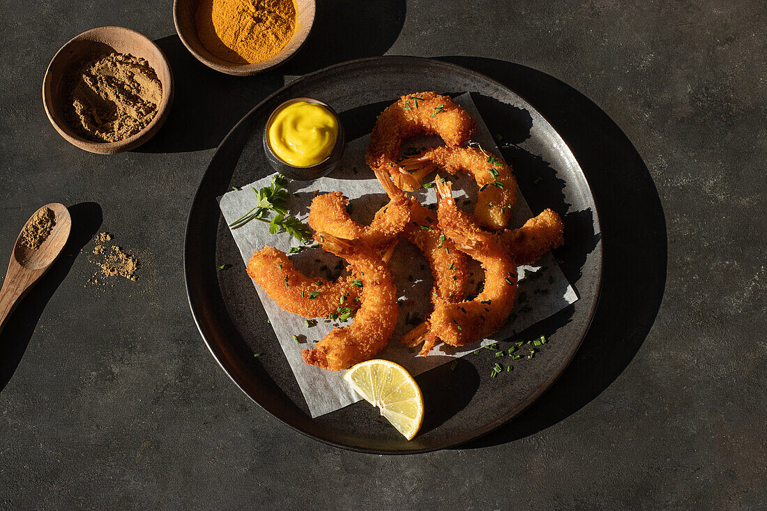 From above still life with plate of lemon and mustard battered prawns with two bowls of turmeric and curry spices