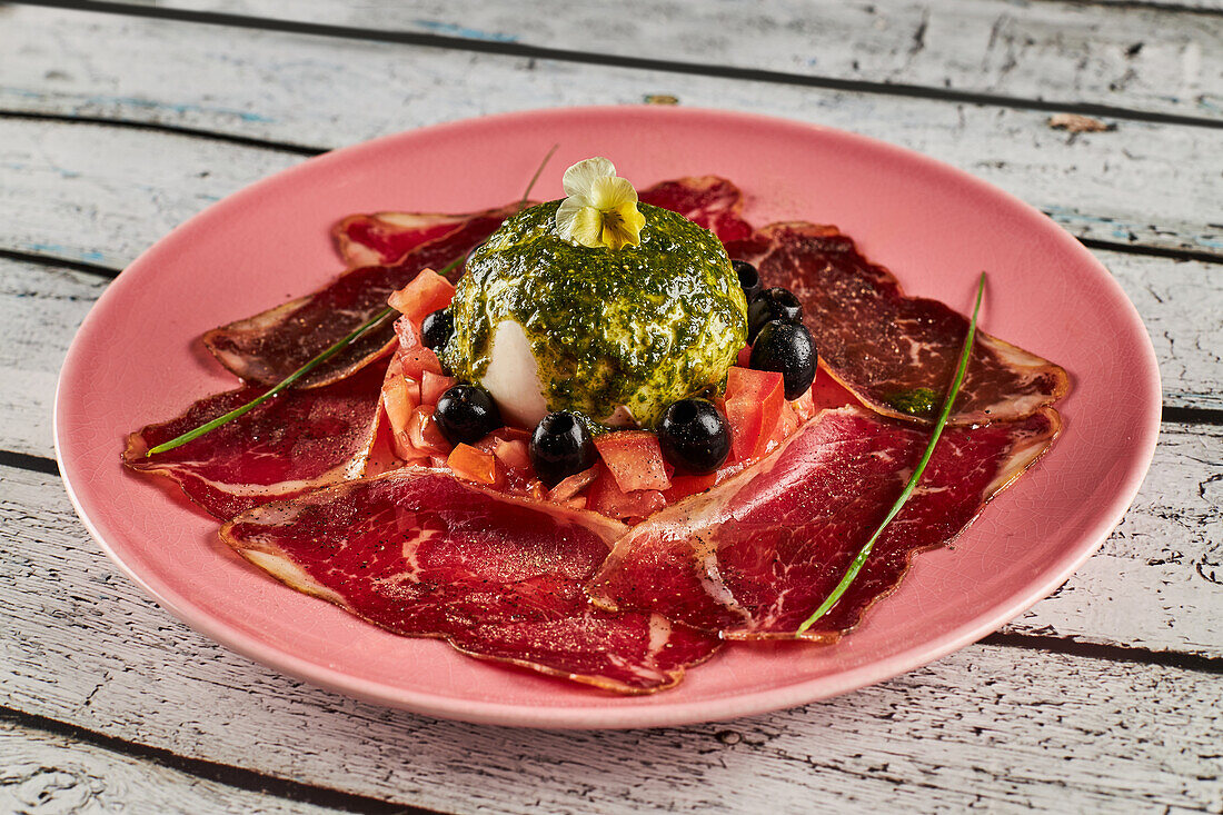 From above tomato salad with burrata and slices of prosciutto garnished with black olives and pesto sauce and served on plate on wooden table