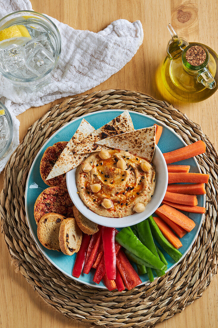 Top view of bowl with hummus and tortilla chips surrounded with various vegetables and croutons served on wicker mat near olive oil and lemonade on timber table
