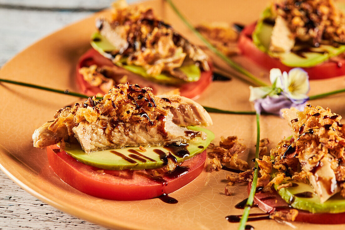 Closeup crispy fried onion and sliced of mackerel placed on slices of tomato and avocado near greens and flowers on plate in restaurant