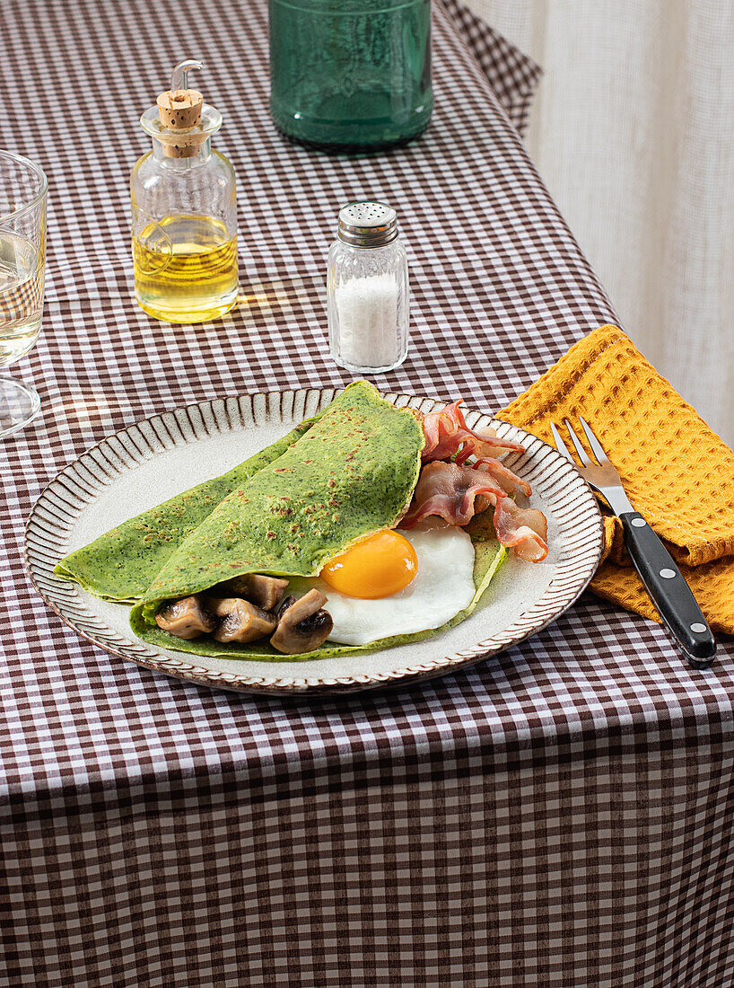 Hausgemachtes Frühstück aus Spinatpfannkuchen mit Speck, Ei und Pilzen, serviert auf einem weißen Teller mit einem Salz- und Ölstreuer auf einer karierten Tischdecke