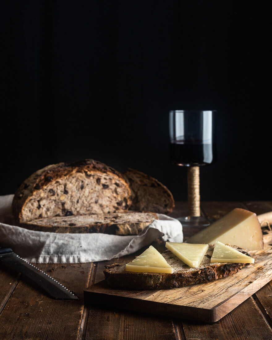 Cut slice of walnut and raisin sourdough bread with loaf on towel served with aromatic cheese and glass of red wine on table