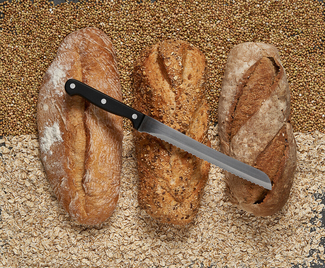 Top view of delicious freshly baked brown loaves with knife placed on raw buckwheat and oat grains in light kitchen
