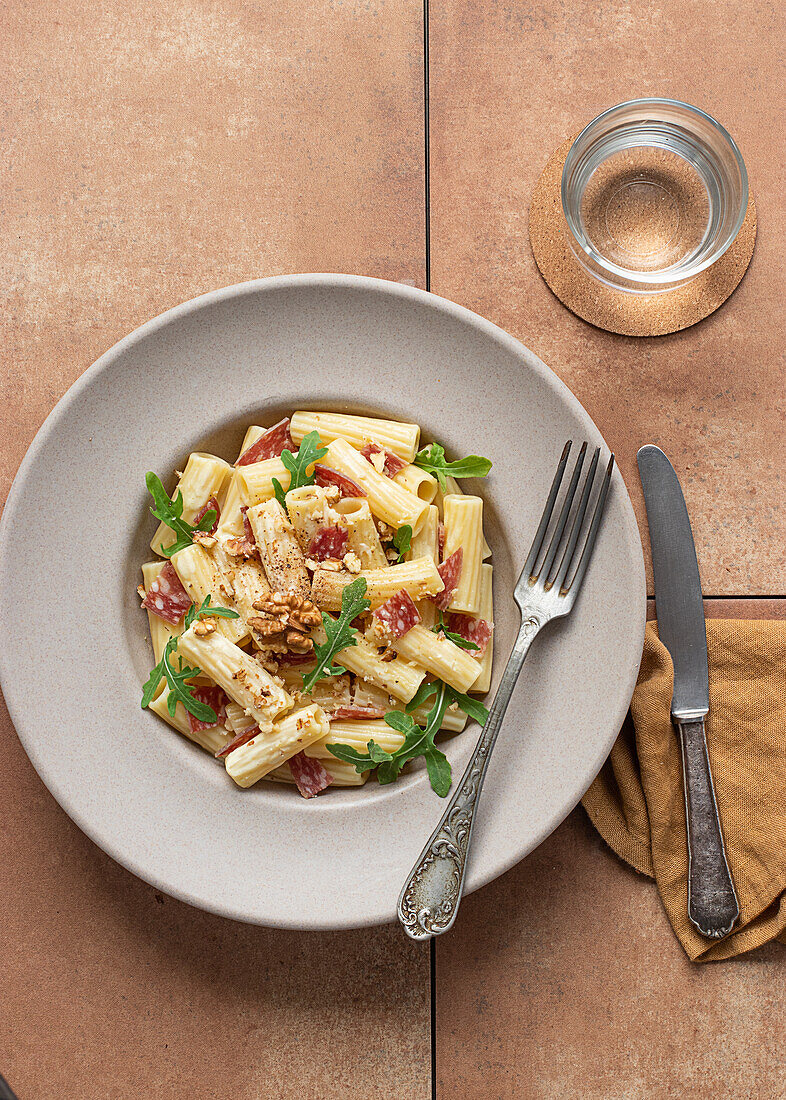 Nudelgericht mit Rucola, Salami und Walnüssen von oben auf einem Tisch, umgeben von Öl, Zitrone und Besteck