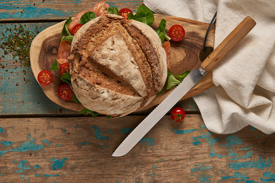 Ansicht von oben von hausgemachtem Sauerteigbrot mit Lachs, serviert auf einem hölzernen Schneidebrett mit Kirschtomaten und Messer auf einem schäbigen Tisch