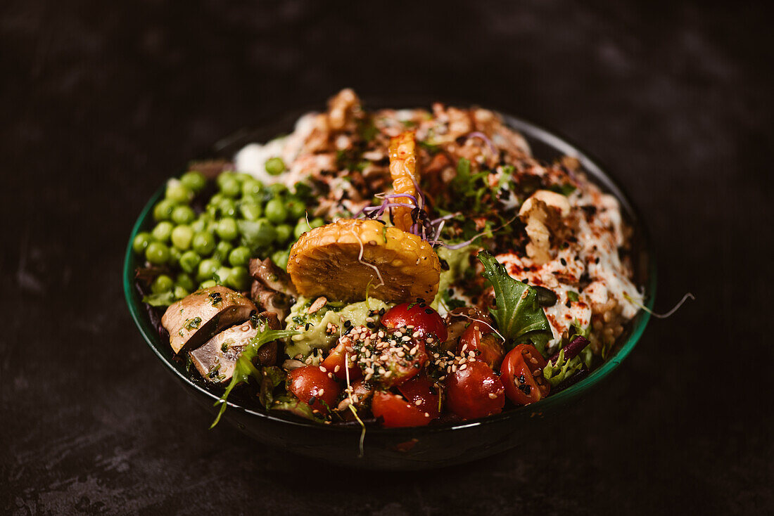 Bowl of tasty sweet potato slices with cherry tomatoes near green peas and sesame seeds on dark background