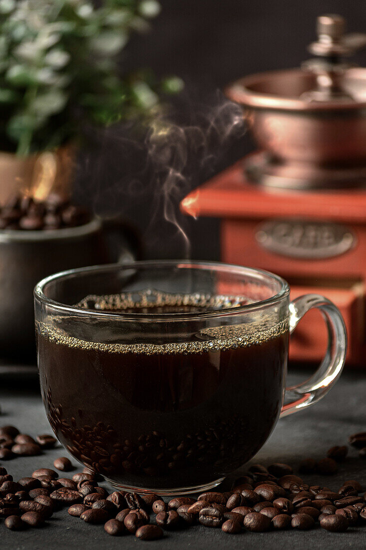 Glass of freshly brewed aromatic coffee on saucer placed on table with scattered beans against manual grinder