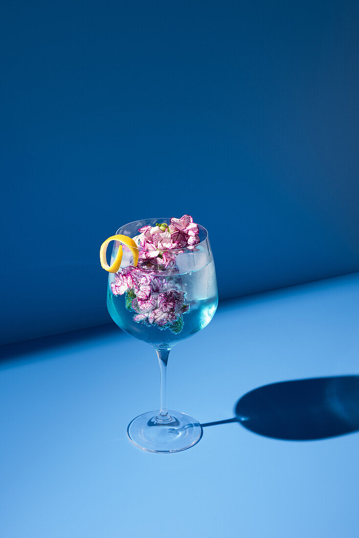 Transparentes Glas mit frischem Cocktail, Minzblättern und Blumen vor blauem Hintergrund auf dem Tisch