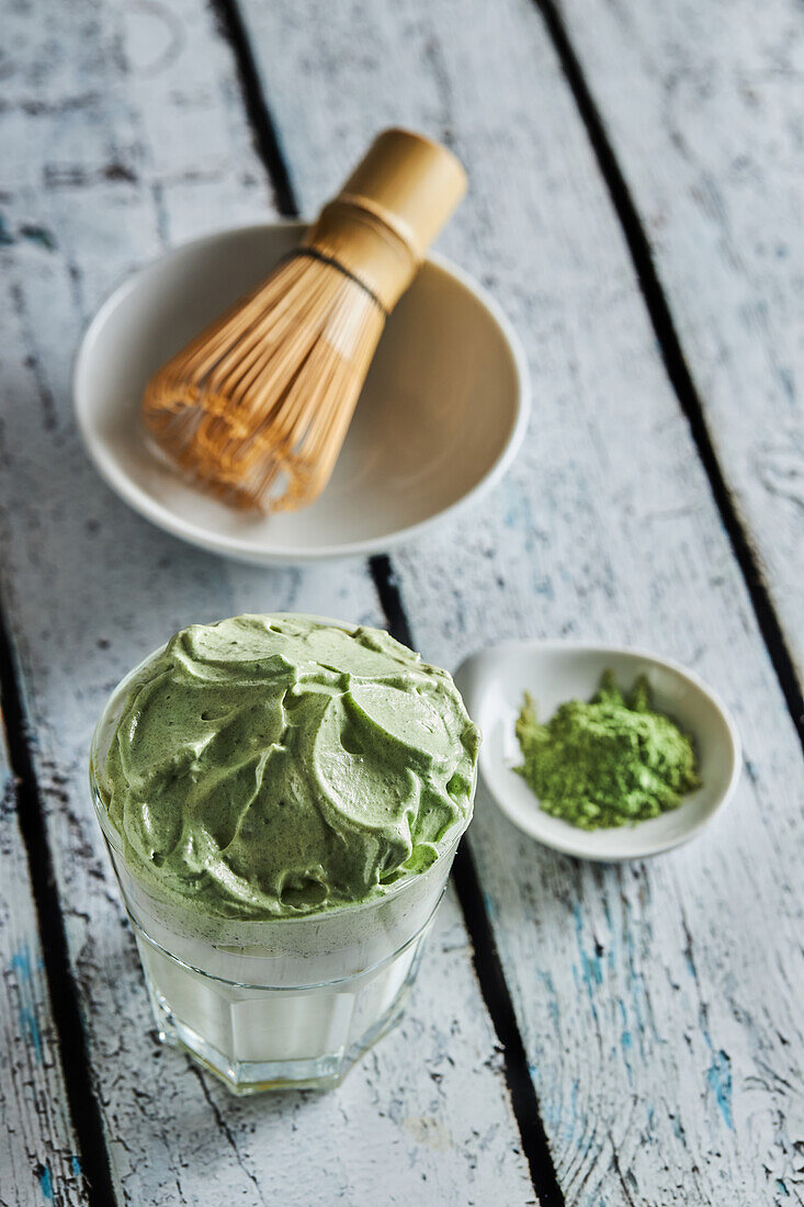 Blick von oben auf ein Glas frischen Dalgona-Kaffee mit Matcha und japanisches Teezeremonie-Zubehör, bestehend aus einem Bambusbesen, genannt Chasen, auf einem Holztisch