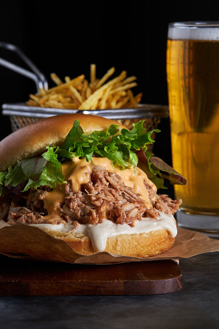 Appetizing burger with pulled pork with barbecue sauce cheddar cheese and lettuce mix served with French fries and glass of cold beer on wooden board