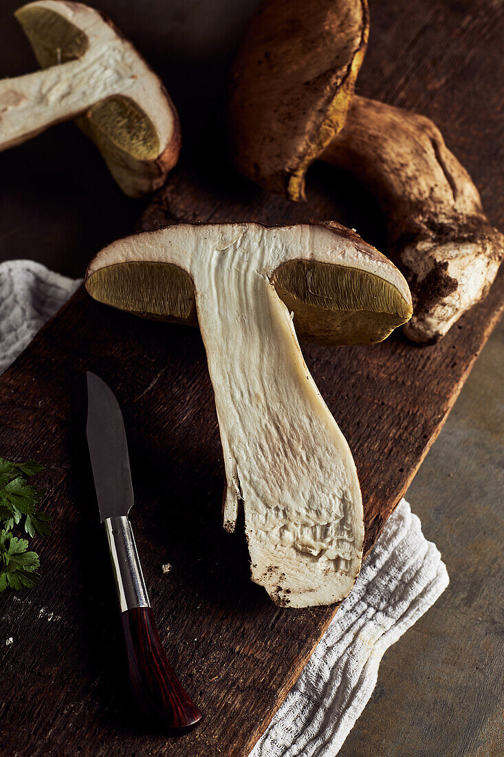 Draufsicht auf roh geschnittene Steinpilze (Boletus edulis) auf hölzernem Schneidebrett mit Knoblauch und Petersilie in heller Küche während des Kochvorgangs