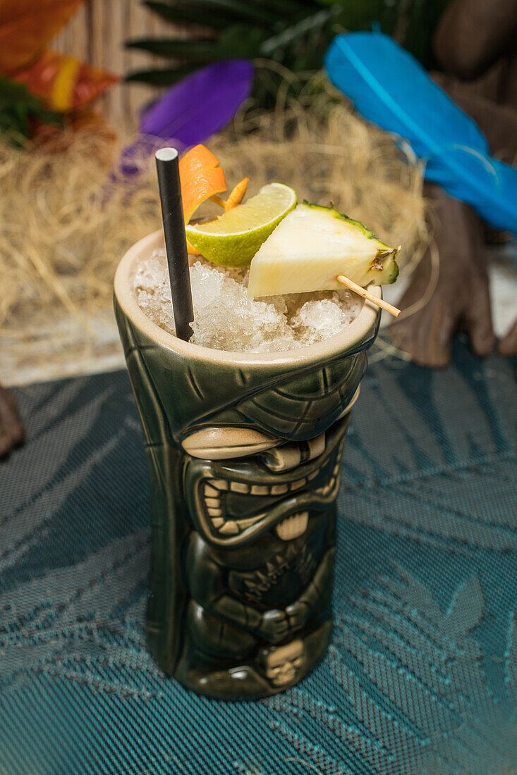 From above of large sculptural tiki cup filled with booze decorated with straw and fruits placed on green rug against dry grass