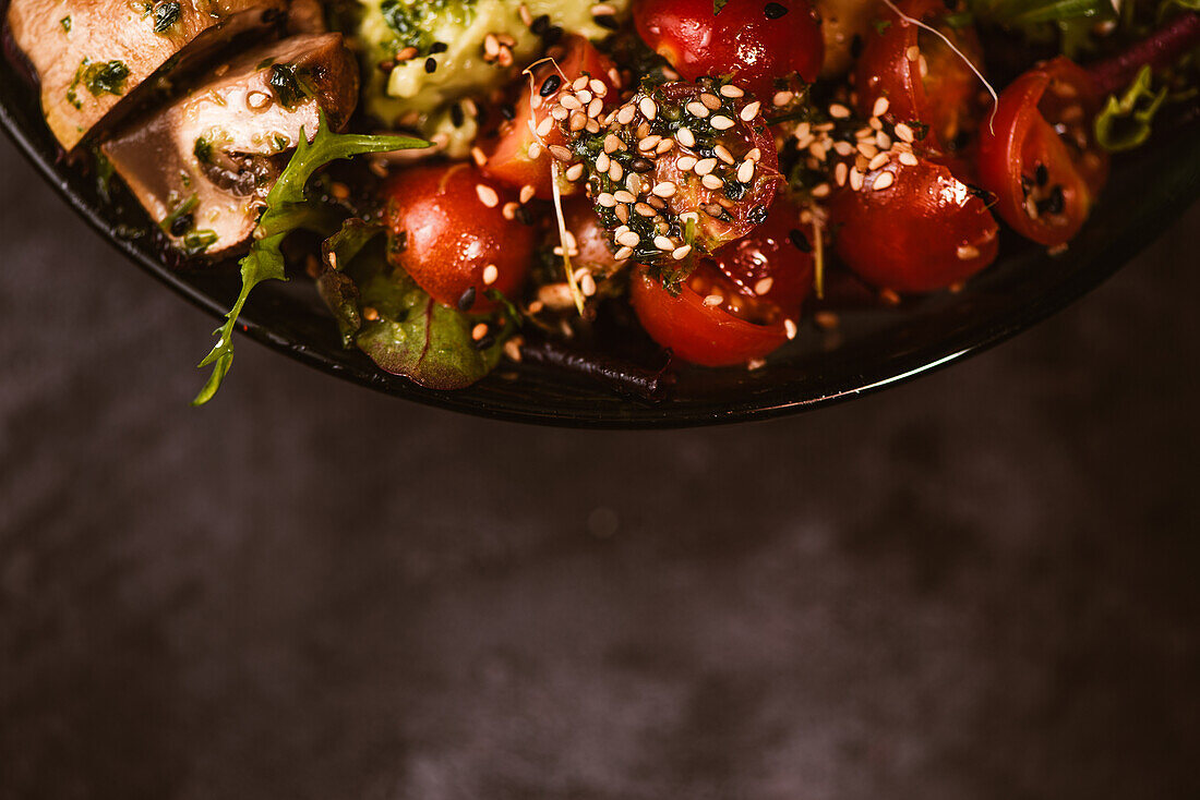 Bowl of tasty sweet potato slices with cherry tomatoes near green peas and sesame seeds on dark background