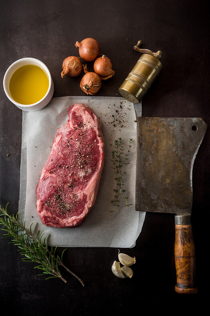 Draufsicht auf ungekochtes Fleischstück mit Gewürzen zwischen frischen Rosmarinzweigen und Knoblauchzehen auf dunklem Hintergrund