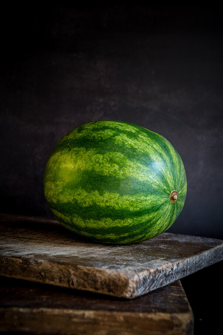 Still life composition of whole ripe striped green watermelon placed on rustic wooden board against black background