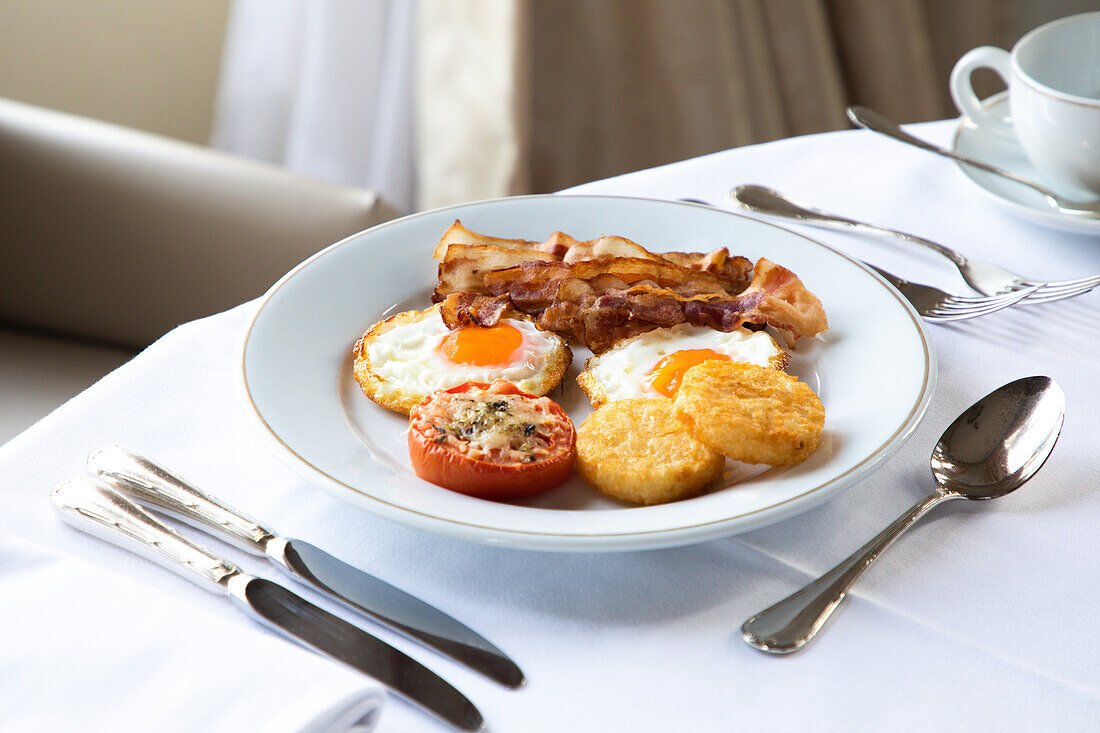 Appetitliche Spiegeleier mit Speckscheiben auf einem Teller mit gefüllter Tomate und knusprigem Mozzarella auf einem Tisch mit Besteck und einer Tasse Kaffee beim Frühstück