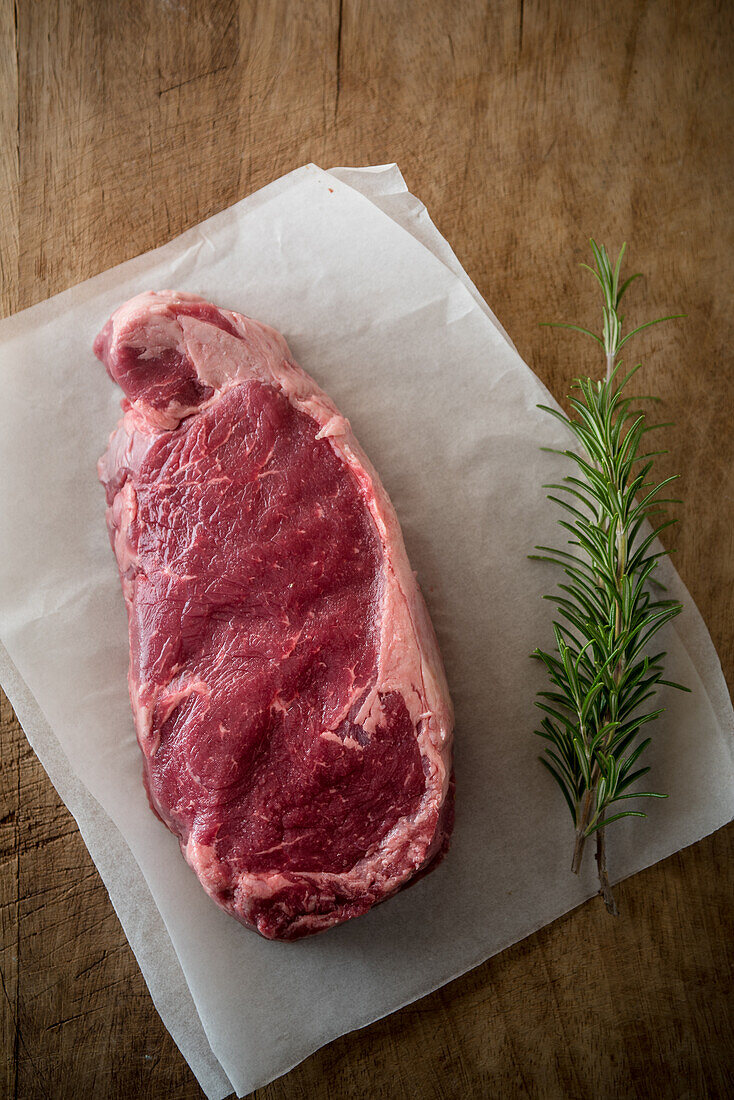 Overhead view of uncooked meat piece with rosemary leaves against baking paper on brown background