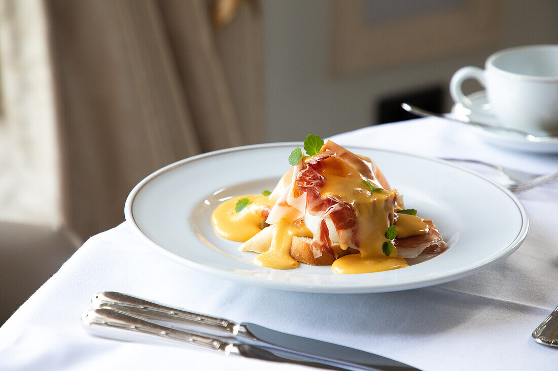 Appetitliche Eier Benedict mit Speck und Sauce Hollandaise, serviert in einem weißen Teller auf einem Tisch mit einer Tasse Kaffee und Besteck in einem Restaurant während des Brunchs