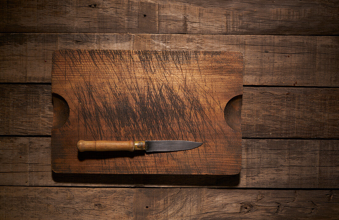 Top view composition with sharp kitchen knife and weathered wooden cutting board placed on rustic plank table
