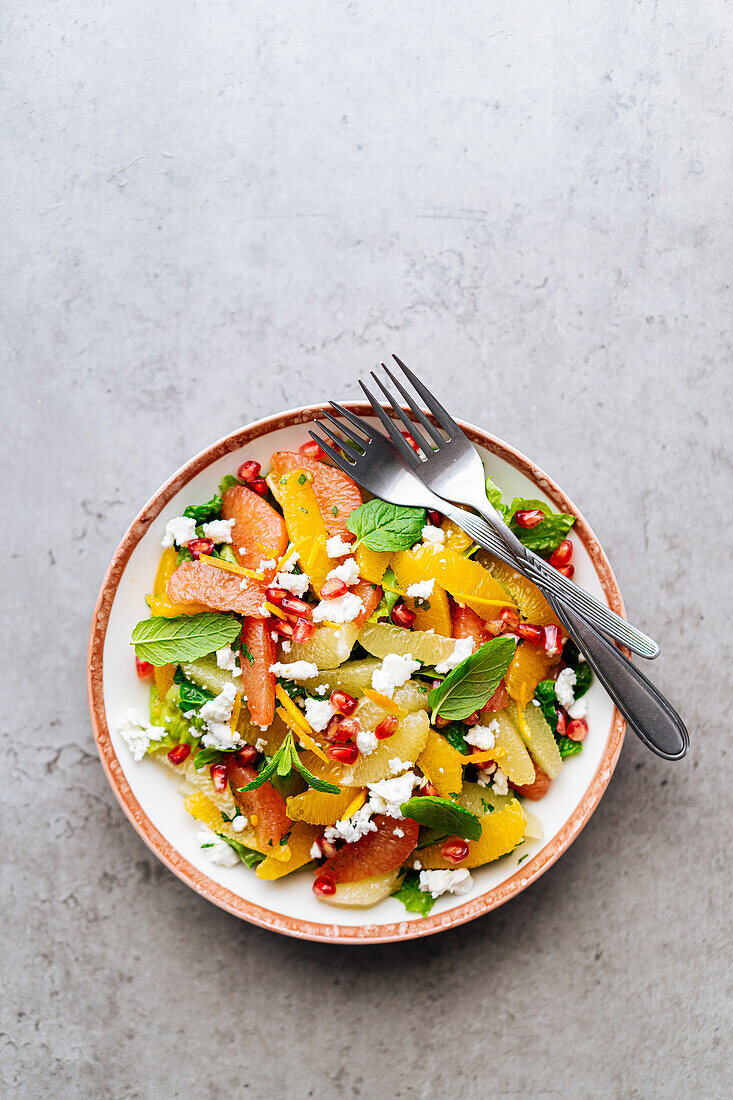 Draufsicht auf traditionellen griechischen Salat mit reifen Tomaten und Feta-Käse, gekrönt von Granatapfelkernen und Minzblättern auf grauem Hintergrund mit Orangensaft
