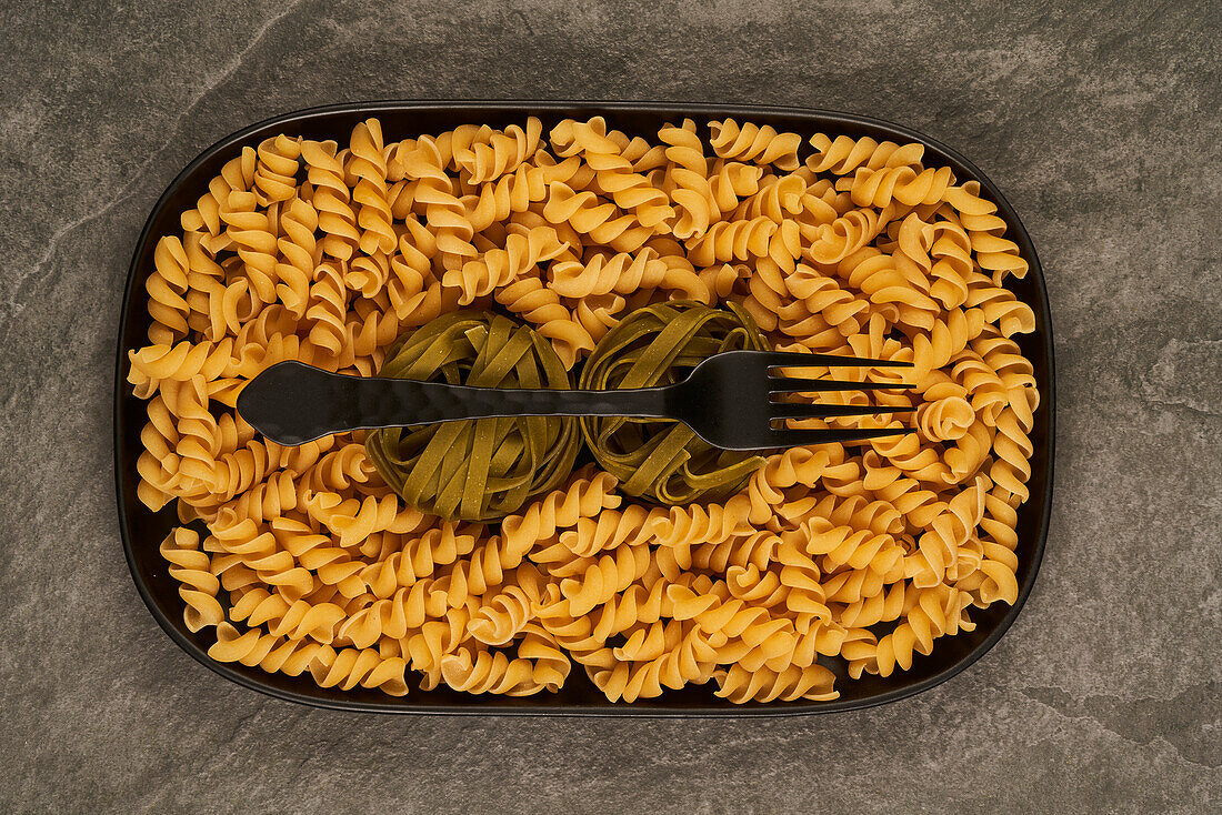 Top view of black fork placed near uncooked fusilli pasta on tray on table